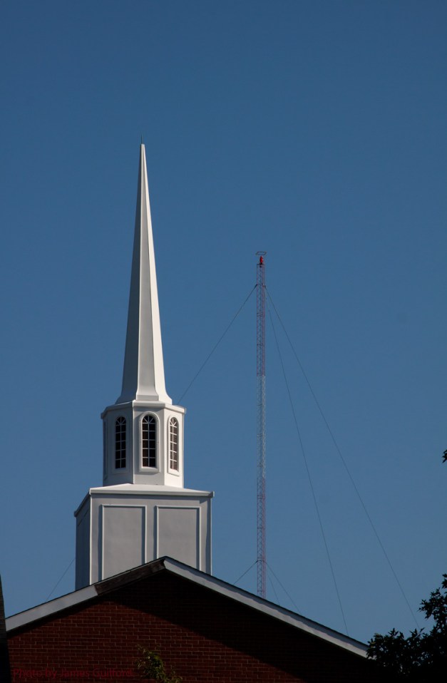 Photo: Church steeple with radio tower in background against a blue sky. Photo by James Guilford.