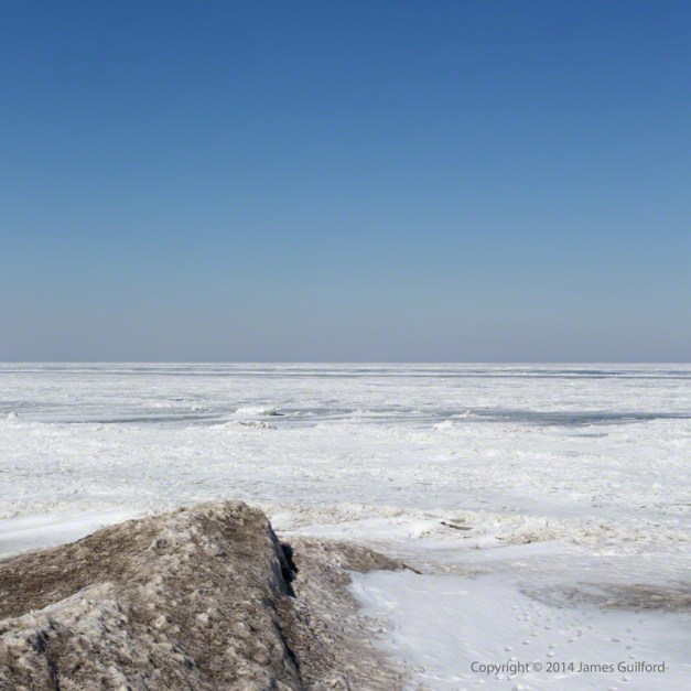 Photo: A spit of sand-tinted ice points northward, out on to the barren expanse of Lake Erie in winter. Photo by James Guilford.