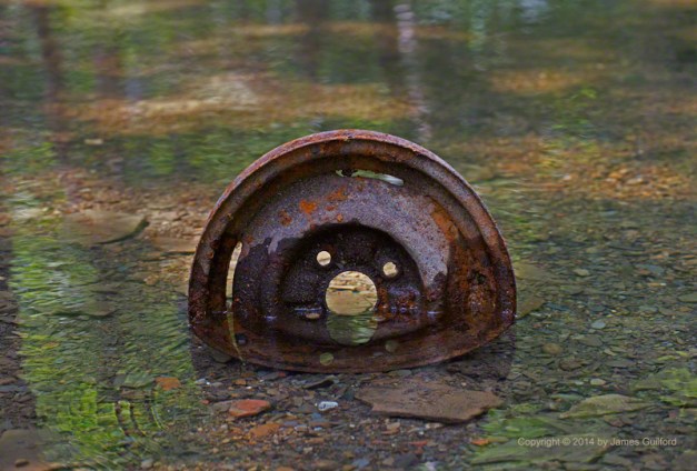 Photo: A rusty automobile wheel sits in a clear running stream. Photo by James Guilford.
