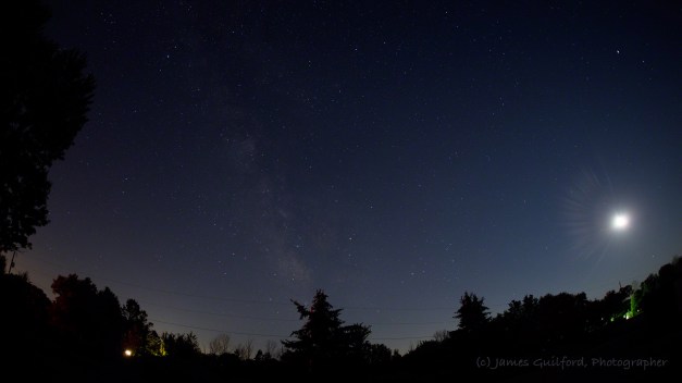 Photo: Wide-angle shot of sky with Milky Way, stars, Moon, and ground visible. Photo by James Guilford.