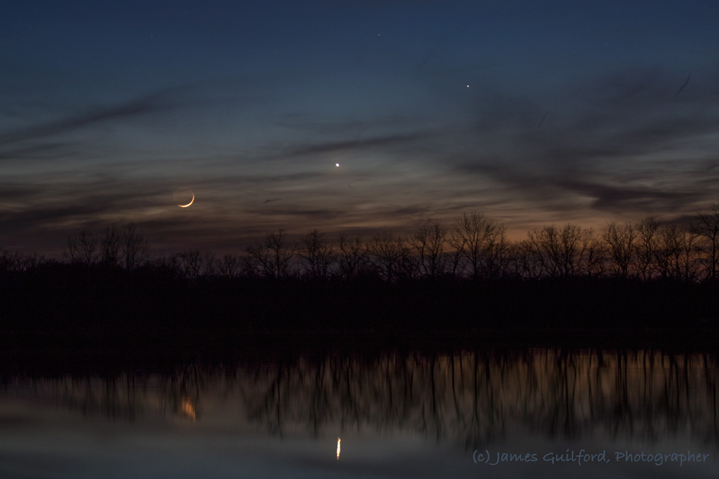 Photo: Parting Shot. The Moon, and Company, sink into clouds and trees to disappear for the night, their light reflected in dark lake waters. Photo by James Guilford.