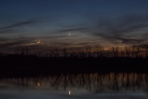 Photo: Parting Shot. The Moon, and Company, sink into clouds and trees to disappear for the night, their light reflected in dark lake waters. Photo by James Guilford.