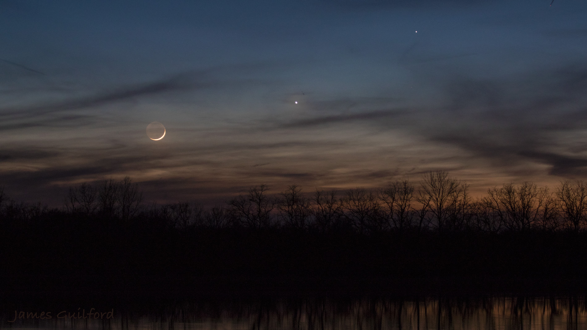 Photo: Colors of Twilight. Left to right: Moon, Venus, and Mercury, in conjunction, sink in the tree-edged western sky. Photo by James Guilford.